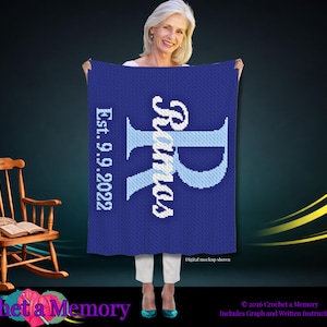 May include: A rectangular blue crochet blanket featuring a large light blue letter R, the name Ramos in white, and the text Est. 9.9.2022 in white. The blanket is displayed against a dark background with a wooden rocking chair visible to the left.