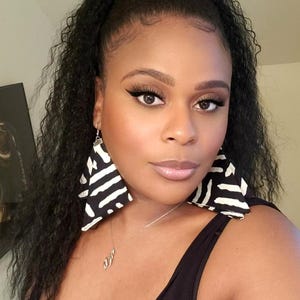 May include: A woman wearing large, triangular earrings with a black and white zebra stripe pattern. She is also wearing a black tank top and a silver necklace. Her dark hair is pulled back in a ponytail.