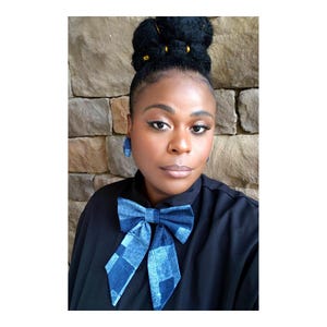 May include: A woman wearing a black top with a denim bow tie and matching earrings. The bow tie has a checkered pattern in shades of blue. The earrings are oval-shaped and also denim.