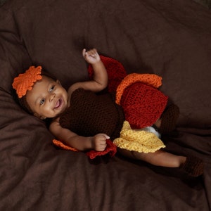 May include: A baby wearing a brown, orange, and yellow crocheted turkey costume. The baby is lying on a brown beanbag chair and smiling at the camera.