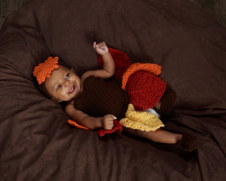 May include: A baby wearing a brown, orange, and yellow crocheted turkey costume. The baby is lying on a brown beanbag chair and smiling at the camera.