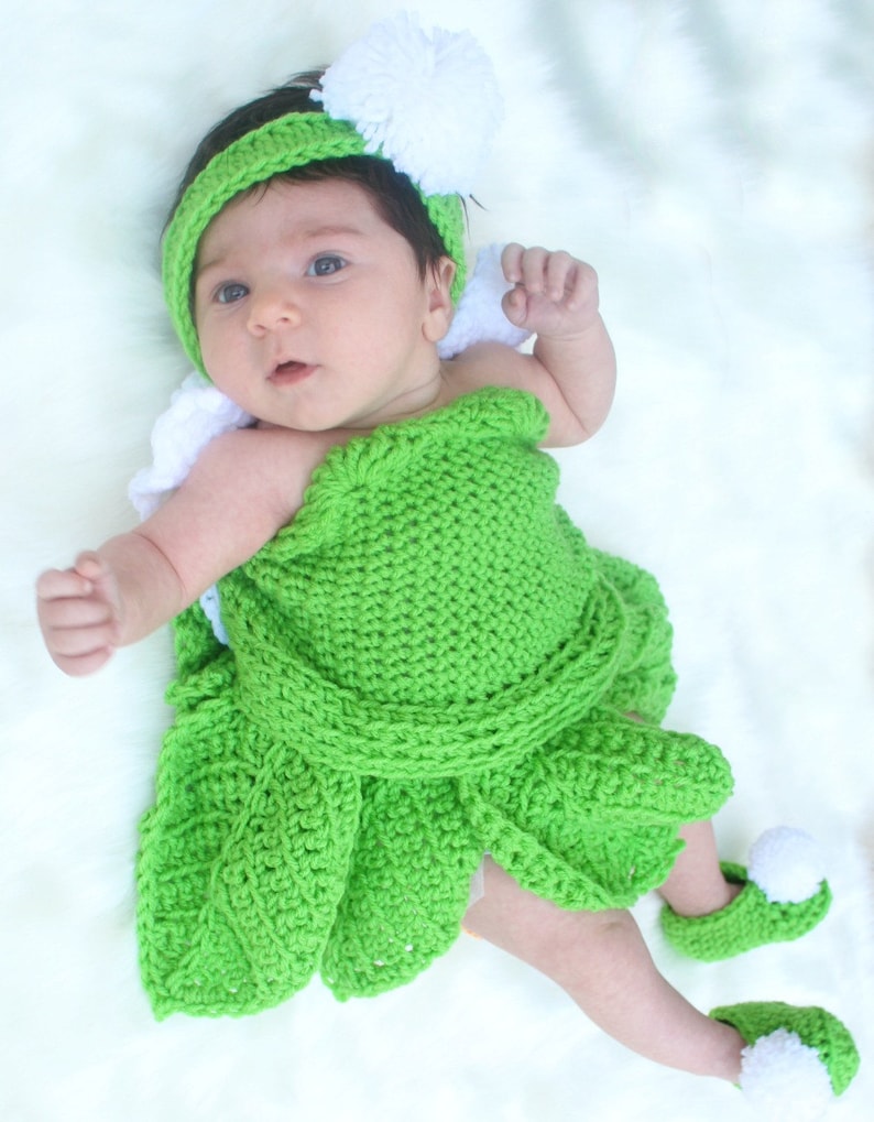 May include: A baby wearing a green crocheted dress with white pom-poms on the hat and shoes.