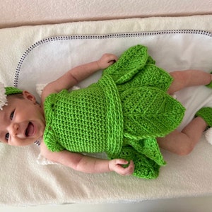 May include: A green crocheted baby dress with leaf details. The dress has a white crocheted headband with a pom-pom.