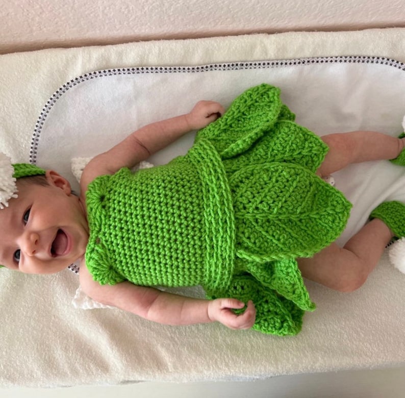 May include: A green crocheted baby dress with leaf details. The dress has a white crocheted headband with a pom-pom.