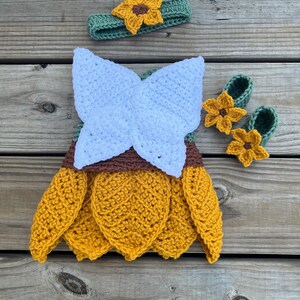 May include: A crocheted sunflower baby outfit with a headband, dress, and booties. The dress is yellow with white wings and brown accents. The headband and booties are green with yellow sunflower accents.