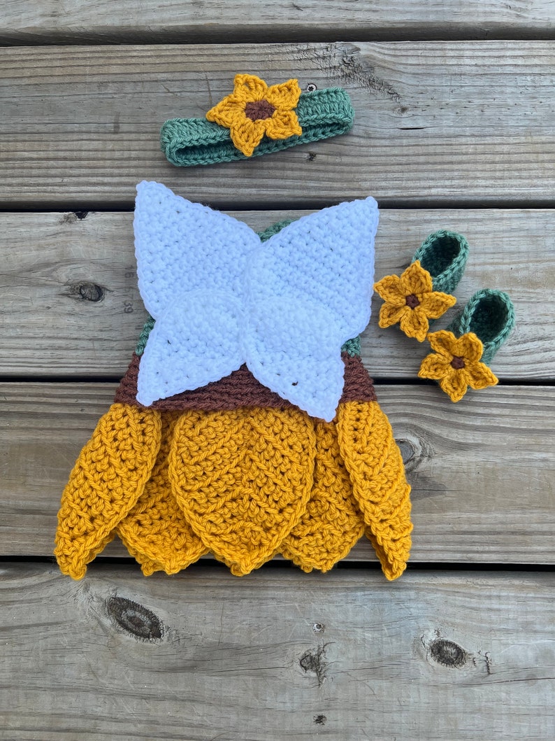 May include: A crocheted sunflower baby outfit with a headband, dress, and booties. The dress is yellow with white wings and brown accents. The headband and booties are green with yellow sunflower accents.
