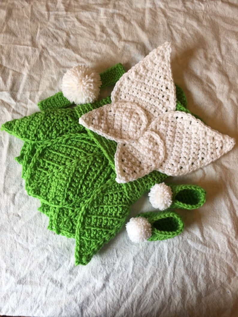 May include: A green crocheted baby outfit with white crocheted wings, a headband, and booties. Each piece has a white pom-pom.