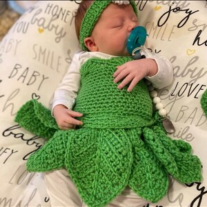 May include: A green crocheted baby dress with a leaf design. The dress has a white crocheted headband and a white beaded pacifier clip.