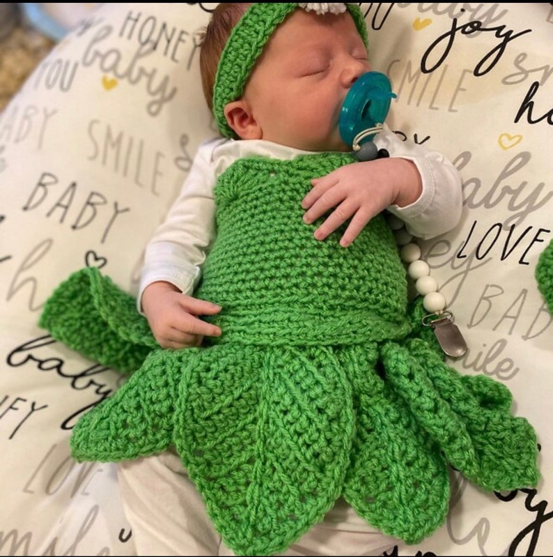 May include: A green crocheted baby dress with a leaf design. The dress has a white crocheted headband and a white beaded pacifier clip.