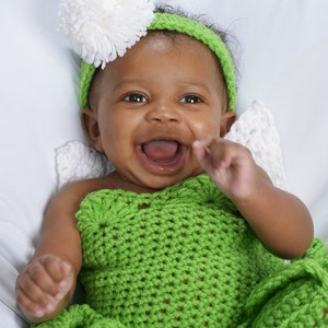May include: A smiling baby wearing a green crocheted outfit with a white pom-pom headband.
