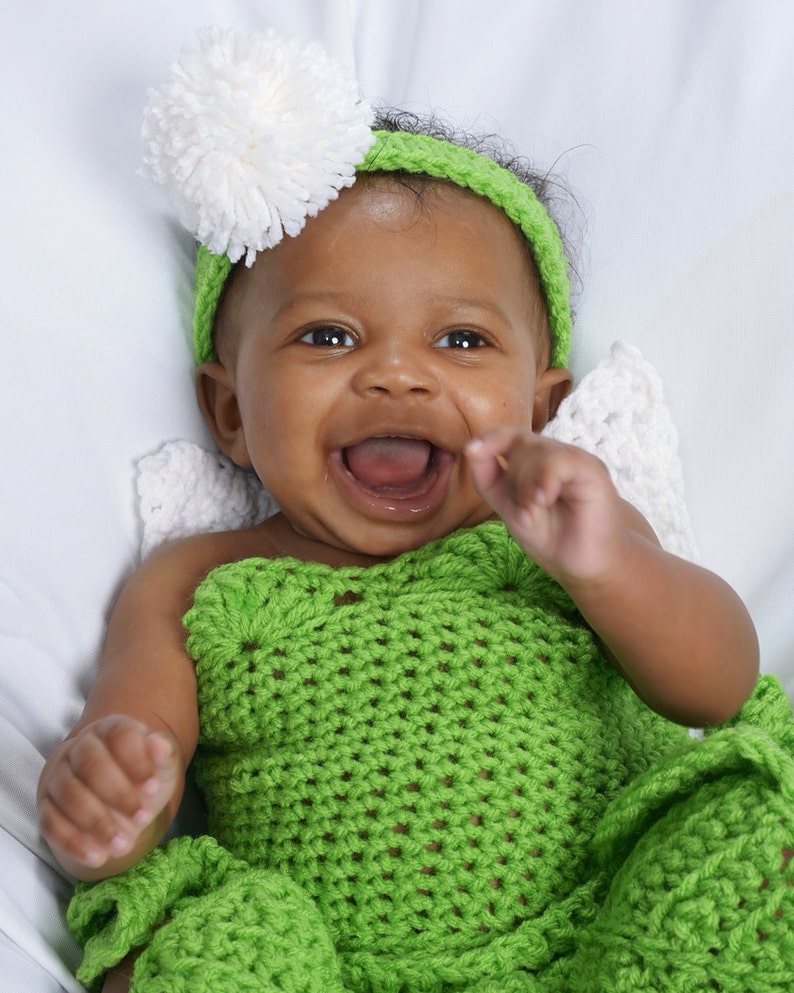May include: A smiling baby wearing a green crocheted outfit with a white pom-pom headband.