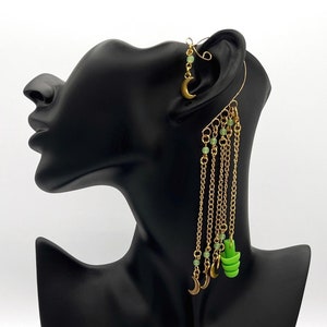 Puede incluir: Un pendiente de aro dorado con cadenas colgantes y abalorios verdes y dorados. Los abalorios incluyen medias lunas y pequeñas cuentas verdes.