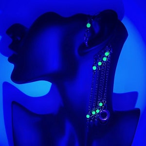 May include: A black mannequin head with a long silver chain earring with green glow-in-the-dark beads. The mannequin is against a blue background.