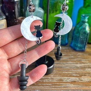 May include: A pair of earrings featuring a black cat silhouette on a white crescent moon charm. The earrings are dangling from a silver chain and have a black earplug charm at the bottom.