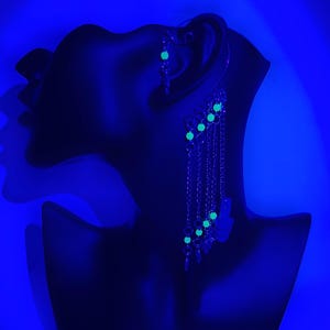 May include: A pair of silver earrings with a dangling chain and green glow-in-the-dark beads. The earrings are on a black mannequin head against a blue background.