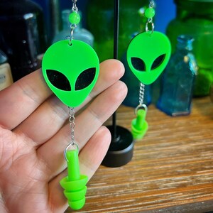 May include: A pair of green alien-shaped earrings with black eyes and a dangling green earplug charm.
