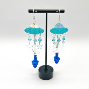 Puede incluir: Un par de pendientes colgantes azules y plateados con un dije de OVNI, dijes de luna y estrellas y tapones para los oídos de silicona azul.