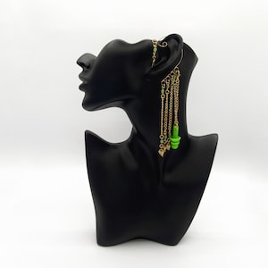 May include: A gold ear cuff with green and gold beads hanging from it. The ear cuff is on a black mannequin head.