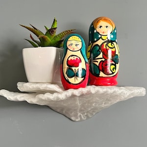 Może przedstawiać: Dwie malowane drewniane lalki matrioszki, jedna czerwona, a druga niebieska, z kwiatowymi wzorami, stoją na białej półce z małą doniczką z sukulentem.