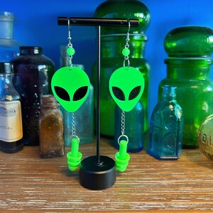May include: A pair of green alien-shaped earrings with black eyes and silver chains. The earrings are hanging from a black stand.