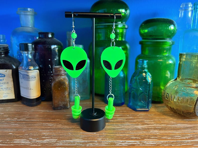 May include: A pair of green alien-shaped earrings with black eyes and silver chains. The earrings are hanging from a black stand.