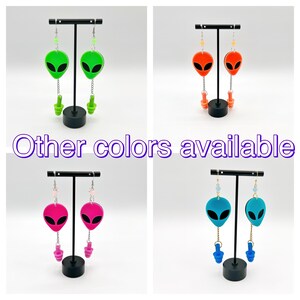 May include: Four pairs of alien-shaped earrings with different colored accents. The earrings are displayed on black stands. The text "Other colors available" is visible in the center of the image.