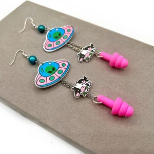 Puede incluir: Pendientes colgantes con gancho plateado, una cuenta azul verdoso, un dije de OVNI rosa y azul con un alienígena verde, un pequeño dije de vaca en blanco y negro y un tapón para los oídos de silicona rosa brillante.