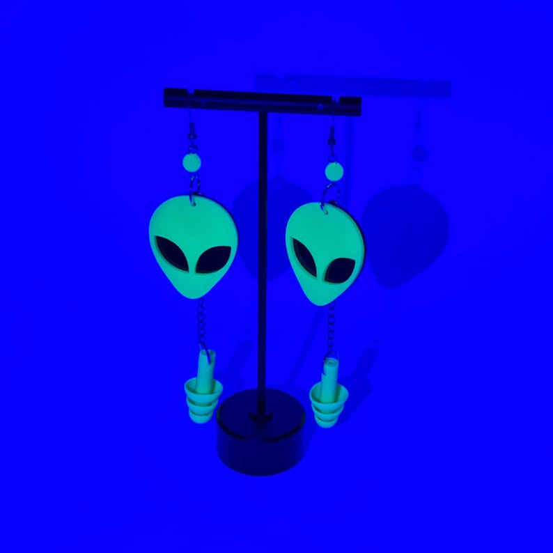 May include: A pair of green alien-shaped dangle earrings with a silver chain and a small green bead at the top. The earrings are hanging from a black stand against a blue background.