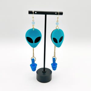 Puede incluir: Un par de pendientes colgantes con un dije de cabeza de alienígena turquesa brillante con ojos negros, una cuenta azul claro y una cadena dorada que conecta con un tapón auditivo de silicona azul. Presentados en un soporte de joyería en forma de T.