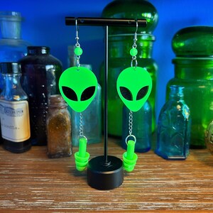May include: A pair of green alien-shaped earrings with black eyes and silver chains. The earrings are hanging from a black stand.