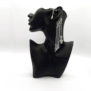 Puede incluir: Pendiente de aro plateado con una cadena colgante adornada con pequeñas cuentas turquesas y abalorios plateados. El pendiente se muestra en una cabeza de maniquí negra.