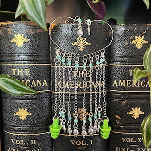 May include: A pair of silver ear cuffs with green beads and green silicone earplugs hanging from them. The ear cuffs are displayed against a background of old books.