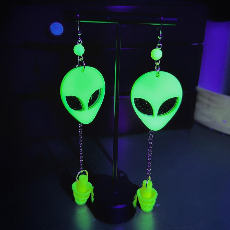 May include: A pair of green glow-in-the-dark alien head earrings with yellow dangling charms. The earrings are hanging from a black stand.