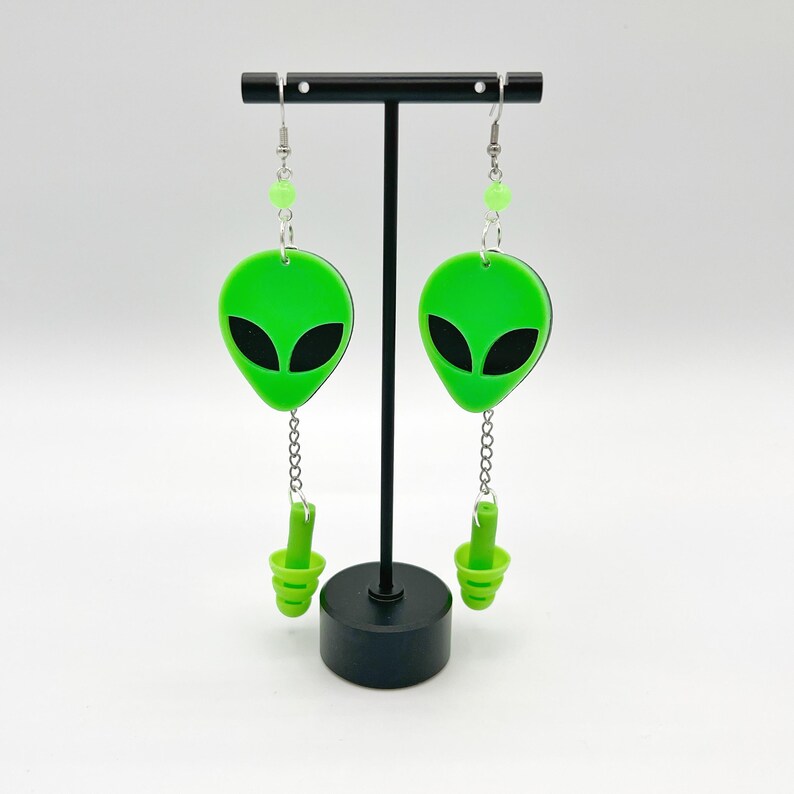 May include: A pair of green alien head dangle earrings with a green dangling charm. The earrings are hanging from a black stand.