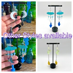 May include: Three pairs of dangle earrings with different designs. The first pair features a crescent moon with a black cat silhouette. The second pair features a blue and white UFO with dangling stars and moons. The third pair features a green UFO with dangling stars and moons.
