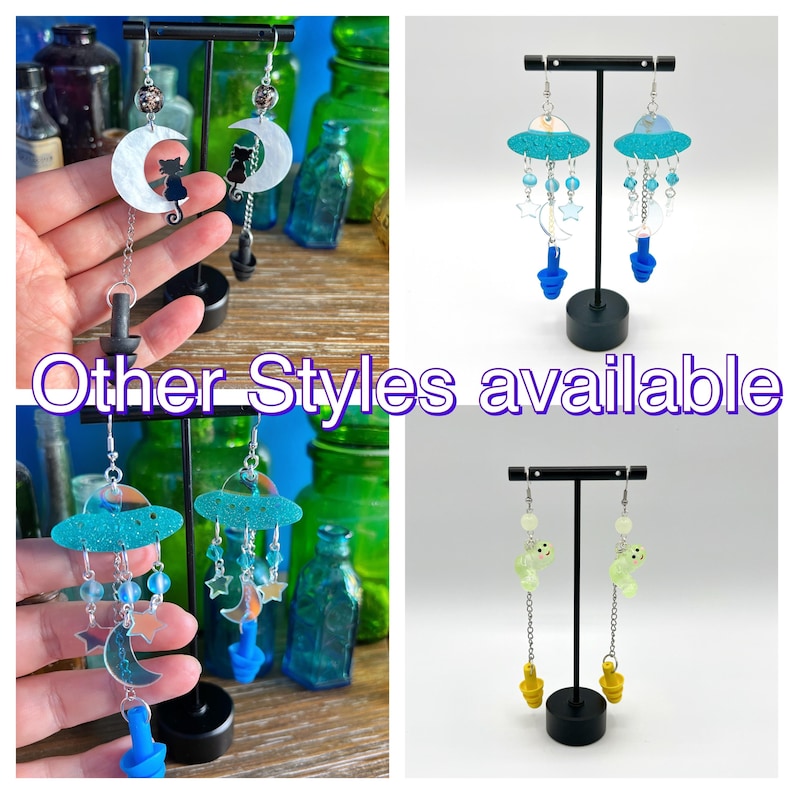 May include: Three pairs of dangle earrings with different designs. The first pair features a crescent moon with a black cat silhouette. The second pair features a blue and white UFO with dangling stars and moons. The third pair features a green UFO with dangling stars and moons.