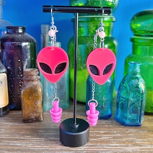 May include: Pink alien head earrings with a dangling pink earplug charm. The earrings are on a black stand.