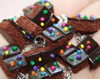Könnte beinhalten: Eine Sammlung von Miniatur-Brownie-Anhängern mit einer texturierten braunen Kuchenbasis, die mit einer glatten dunklen Schokoladenschicht und bunten Zuckerstreuseln verziert ist. Jeder Anhänger hat einen silbernen Metallring zur Befestigung.