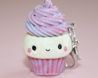 Könnte beinhalten: Ein kleiner, weißer Cupcake mit rosa und lila Swirl-Frosting und einem fröhlichen Gesicht. Der Cupcake ist mit rosa und lila Streifen verziert und hat einen silbernen Anhänger.