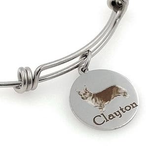 May include: Silver bangle bracelet with a circular charm. The charm displays a brown and white corgi illustration and the name "Clayton". The bracelet has a simple, adjustable design.