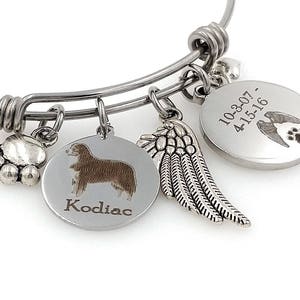 Może przedstawiać: Srebrna bransoletka z zawieszkami z wieloma wisiorkami. Zawieszki obejmują odcisk łapy, sylwetkę psa z imieniem "Kodiac", skrzydło oraz okrągłą zawieszkę z datami i wzorem odcisku łapy i skrzydła.