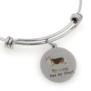 May include: A silver bangle bracelet with a circular charm. The charm displays a brown and white corgi illustration and the text "My corgi has my heart". The bracelet has a simple, elegant design.