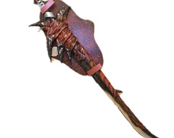 May include: A detailed, fantasy-themed sword with a brown and red handle and blade. The sword is mounted on a brown plaque with pink straps. The blade has a curved design with a sharp edge, and the handle has intricate details.