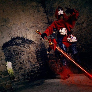 Può includere: Una persona in un costume rosso con cappuccio e una maschera di teschio tiene una spada rossa incandescente in una stanza buia di pietra.