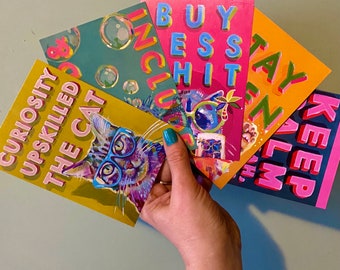 May include: A hand holds five colorful art prints. The prints feature text like "Curiosity Upskilled The Cat" and "Buy Less Shit." Each print has a unique design, including a cat wearing glasses and bubble illustrations. The background is a light teal.