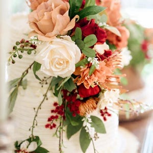 Puede incluir: Un pastel blanco de varios niveles con glaseado de textura horizontal, decorado con un arreglo de flores color melocot&oacute;n, crema y rojo oscuro, follaje verde y peque&ntilde;as bayas rojas que caen por el costado sobre una base de madera.