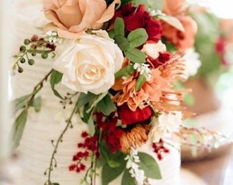 Puede incluir: Un pastel blanco de varios pisos adornado con flores de color melocotón, rojo y naranja. El pastel se coloca sobre un soporte de madera, creando un aspecto rústico y elegante. Hojas verdes y pequeñas flores blancas complementan el arreglo floral.