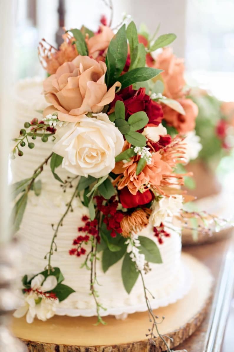 Puede incluir: Un pastel blanco de varios niveles con glaseado de textura horizontal, decorado con un arreglo de flores color melocot&oacute;n, crema y rojo oscuro, follaje verde y peque&ntilde;as bayas rojas que caen por el costado sobre una base de madera.