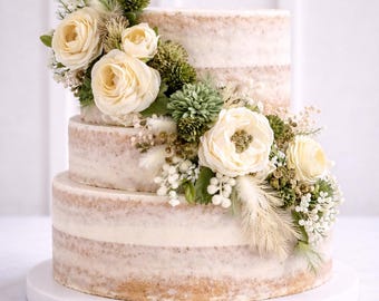 Puede incluir: Un pastel de tres niveles con un diseño rústico, decorado con rosas color crema, acentos verdes y delicadas flores blancas. El pastel se encuentra sobre un soporte blanco, perfecto para una boda o celebración.