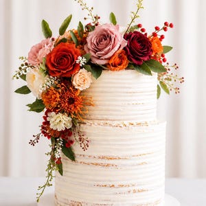 Puede incluir: Un pastel de dos niveles semi-desnudo con glaseado blanco de textura horizontal. La parte superior est&aacute; adornada con un vibrante arreglo floral de rosas en tonos rojo oscuro, rosa polvoriento y naranja, con hojas verdes y bayas rojas.